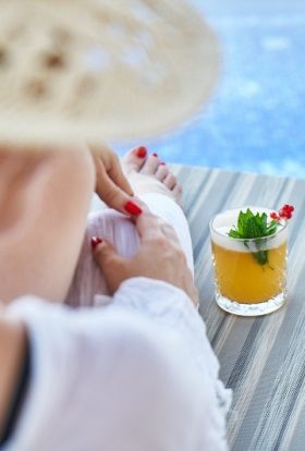 Immune Boosting Cocktail Recipes Photo Overlooking a Woman's Shoulder Sitting on a Beach Chair by the Pool with a Cocktail on the End of the Chair